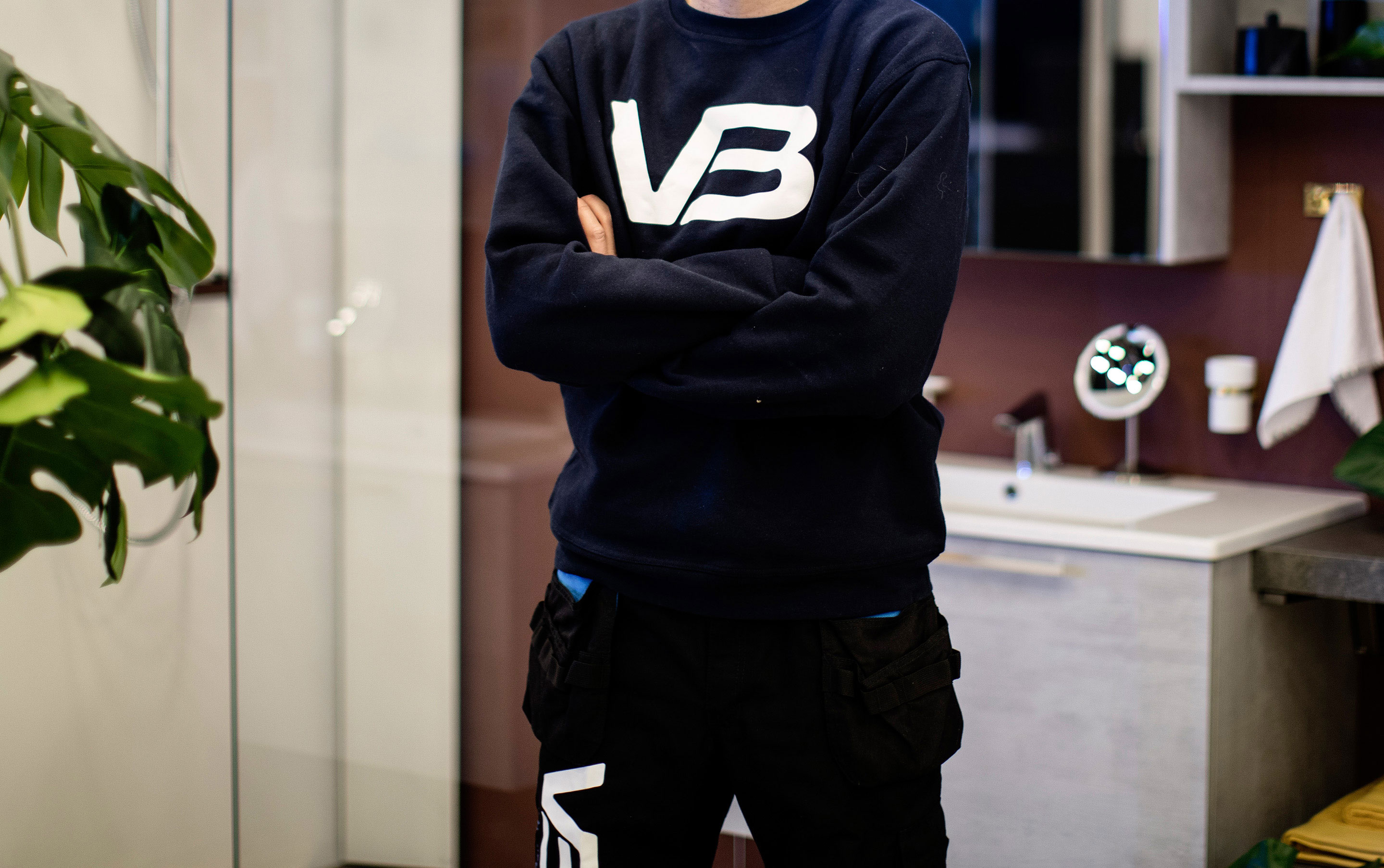 VB-rørlegger i mørkt arbeidsantrekk med armene i kors på baderom med servantskap i grått og hvit porselensvask mot burgunder vegg.