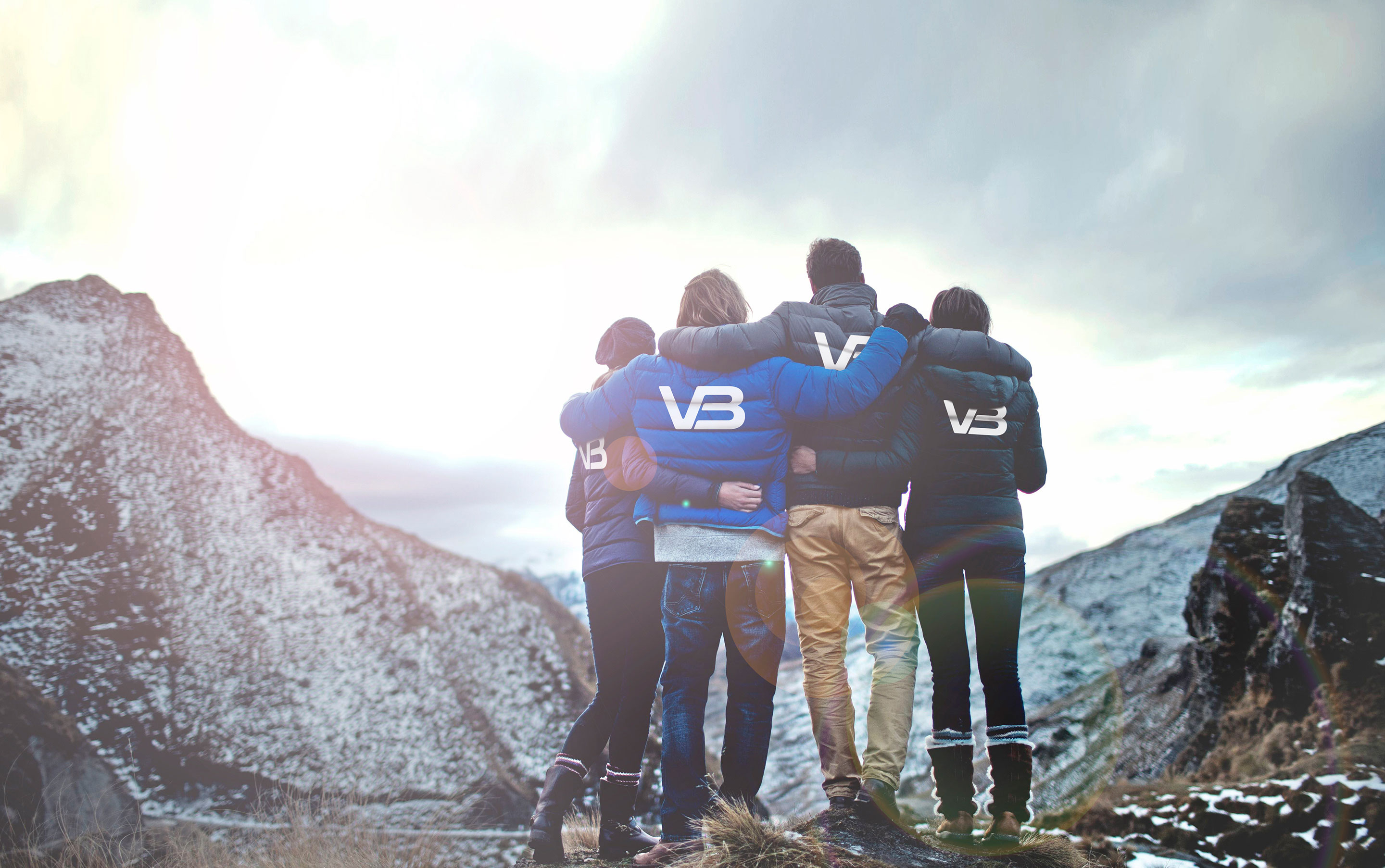 Fire mennesker i boblejakker med VB-logo med armene rundt hverandre og med ryggen til, vendt mot et fjellandskap i disig sollys.